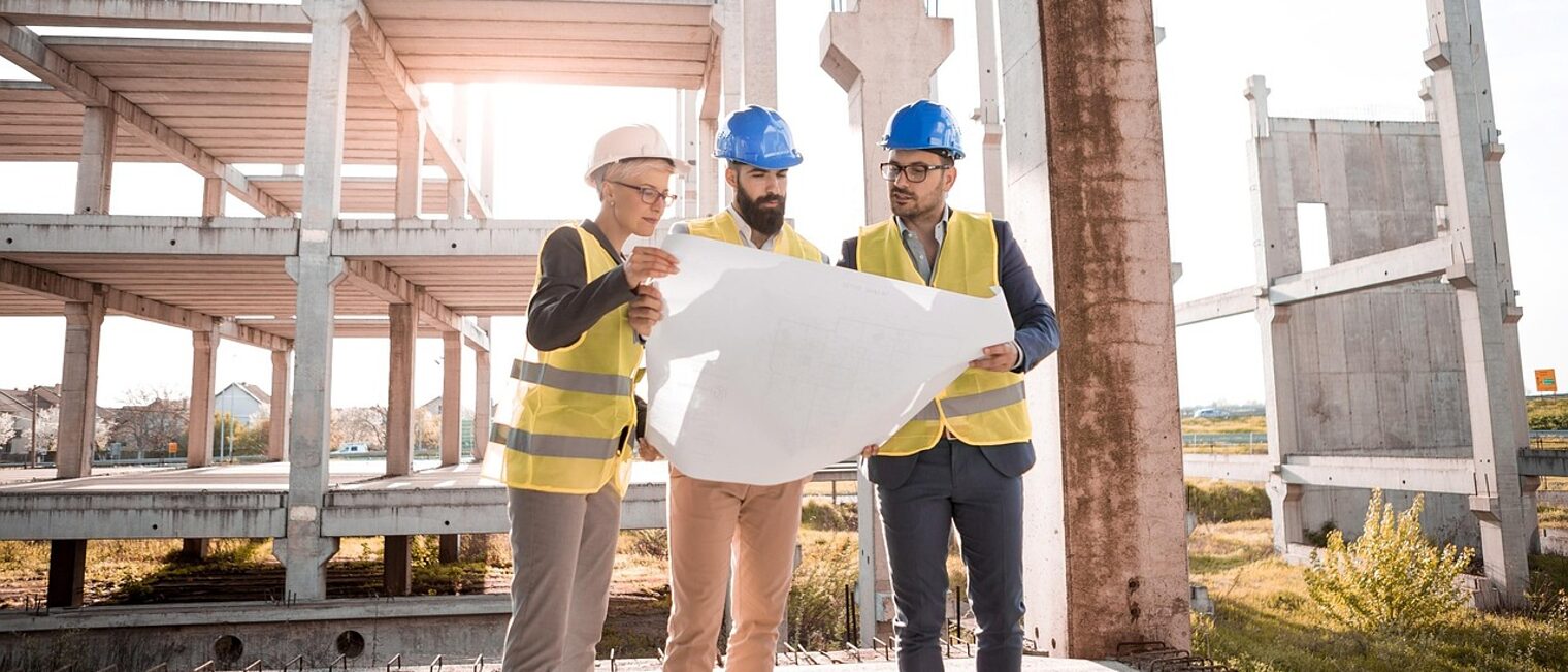 Besprechung Im Hintergrund befindet sich eine Baustelle eines Gebäudes, dass sich im Rohbau befindet. Im Vordergrund stehen drei Männer mit Warnerste und Helm, die alle auf einen Bauplan gucken.