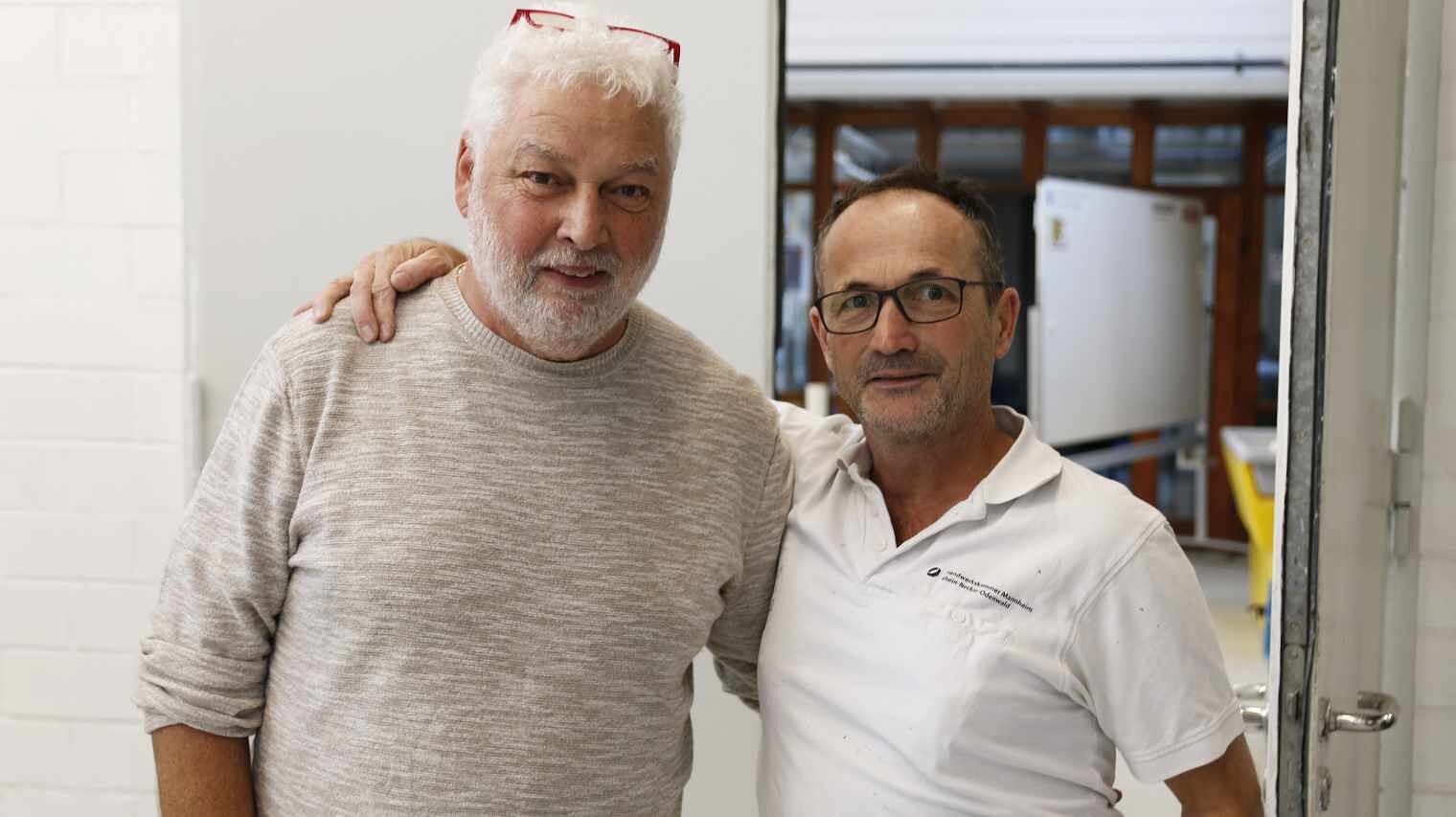 Zwei Männer schauen in die Kamera, der linke etwas größer mit weißem Haar und weißem Vollbart und roter Brille auf dem Kopf, der rechte klein mit lichtem dunklem Haar und weißem Maler-Shirt.