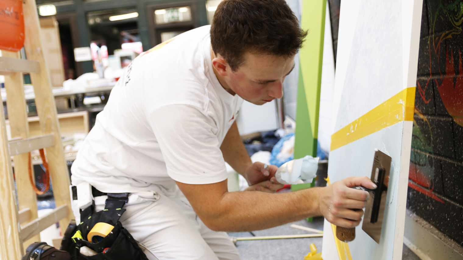 Junger Mann in Malerkleidung kniet vor einer Wand und beugt sich nach vorne, um diese mit dem Spachtel zu bearbeiten inmitten einer Werkstatt-Umgebung