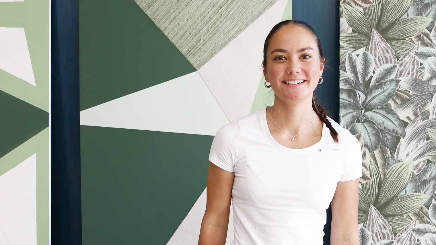 Junge Frau mit Creolen, dunklem zum Zopf geflochtenem Haar, das auf die rechte Schulter fällt und weißem Shirt vor einer mit grünen Flächen bemalen Wand und Fototapete