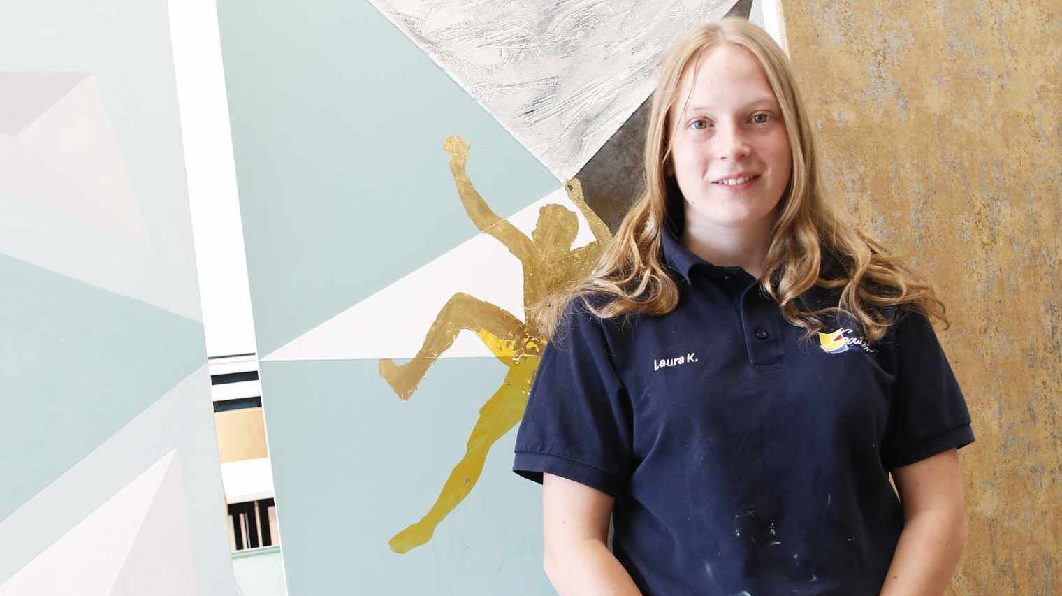 Junge Frau mit blondem, offenem Haar, das auf die Schultern fällt und dunkelblauem Maler-Shirt vor einer Wand mit grafischen Bemalungen und skizziertem Kletterer, lächelt in die Kamera
