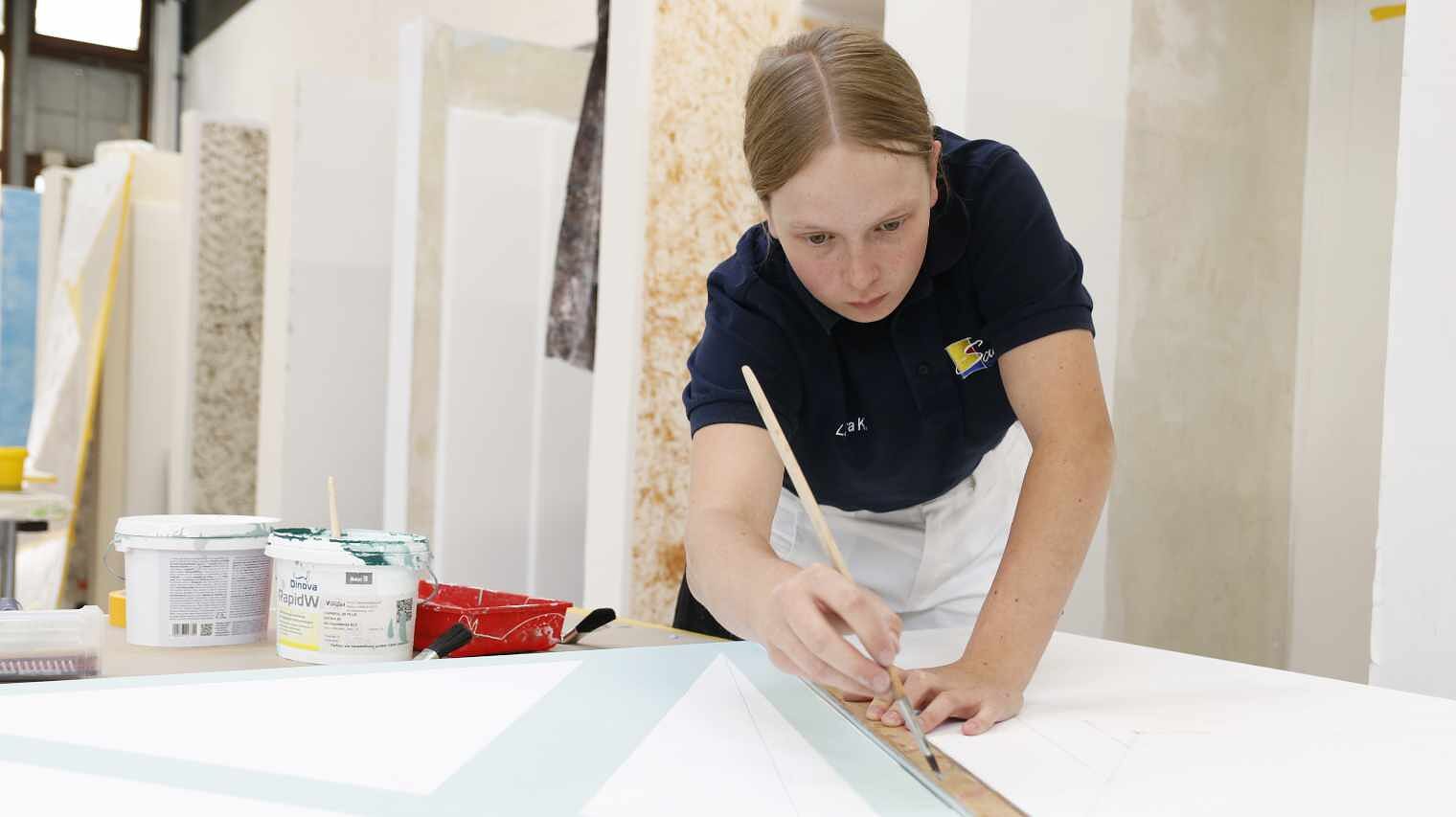 Junge Auszubildende mit zusammengebundenem hellem Haar beugt sich über eine Fläche und zeichnet mit Linial und Stift eine Linie auf, im Hintergrund Maler-Wände