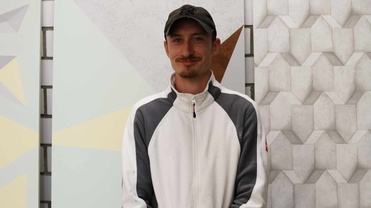 Junger Mann mit grauem Käppi, weißem Fließshirt und grauen Streifen an den längen Ärmeln, Schnäuzer und Kinnbart lächelt in die Kamera, dahinter Malerwände