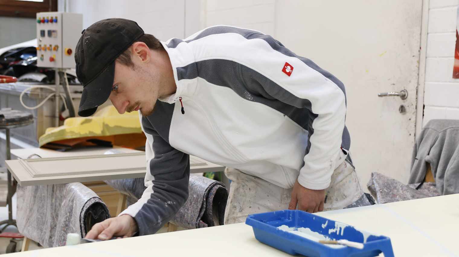 Junger Maler mit grauem Käppi und Maler-Kleidung beugt sich über die Kante einer Arbeitsplatte und streicht mit der Malerrolle darüber, auf der Platte ein blaues Behältnis mit Farbe