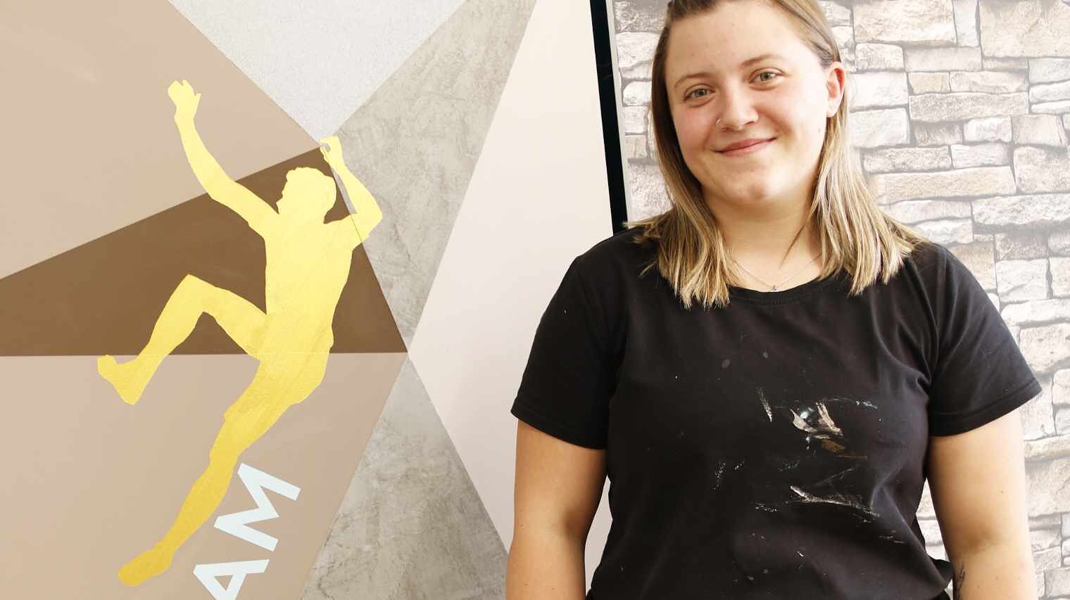 Junge Frau mit schulterlangen blonden Haaren und schwarzem T-Shirt neben einer bemalten Wand mit Kletterer-Motiv lächelt freundlich in die Kamera
