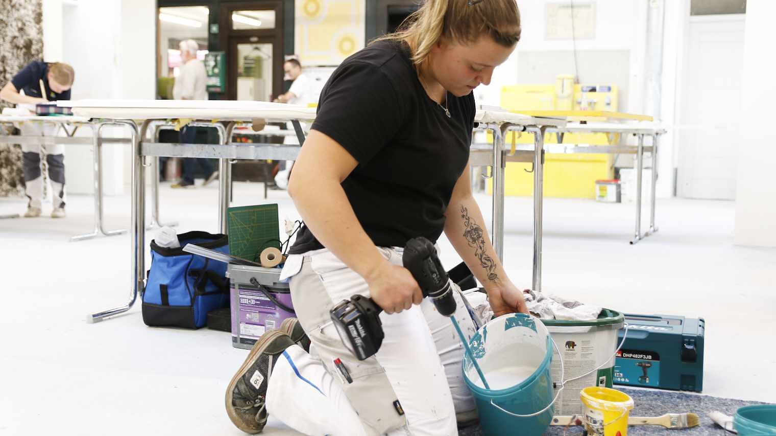 Junge Frau in einer Maler-Werkstatt kniet auf dem Boden und hält einen Farbrührer in einen kleinen Eimer, voll mit Farbe, im Hintergrund Malertische und weitere junge Leute, die arbeiten