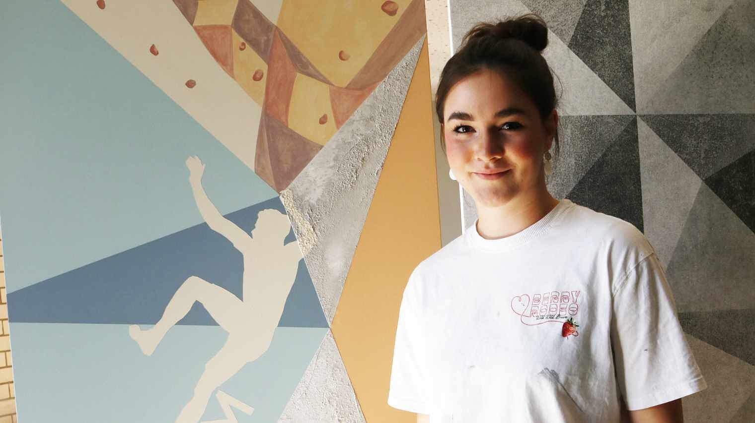 Junge Frau im weißen T-Shirt mit zu einem lockeren Dutt hochgestecktem dunklem Haar neben einer grafisch bemalten Wand mit Kletterer-Motiv, lächelt in die Kamera