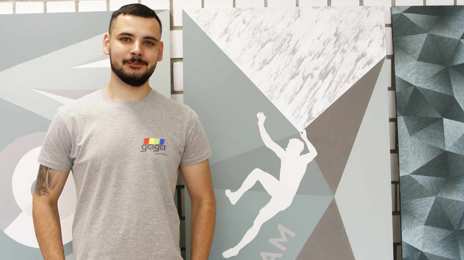 Junger Mann mit sehr kurzem, dunklem Haar und kurzem Vollbart, lichtgraues T-Shirt und Tattoo am linken Oberarm lächelt in die Kamera, hinter ihm grafisch bemalte Wände, eine mit Ketterer-Motiv