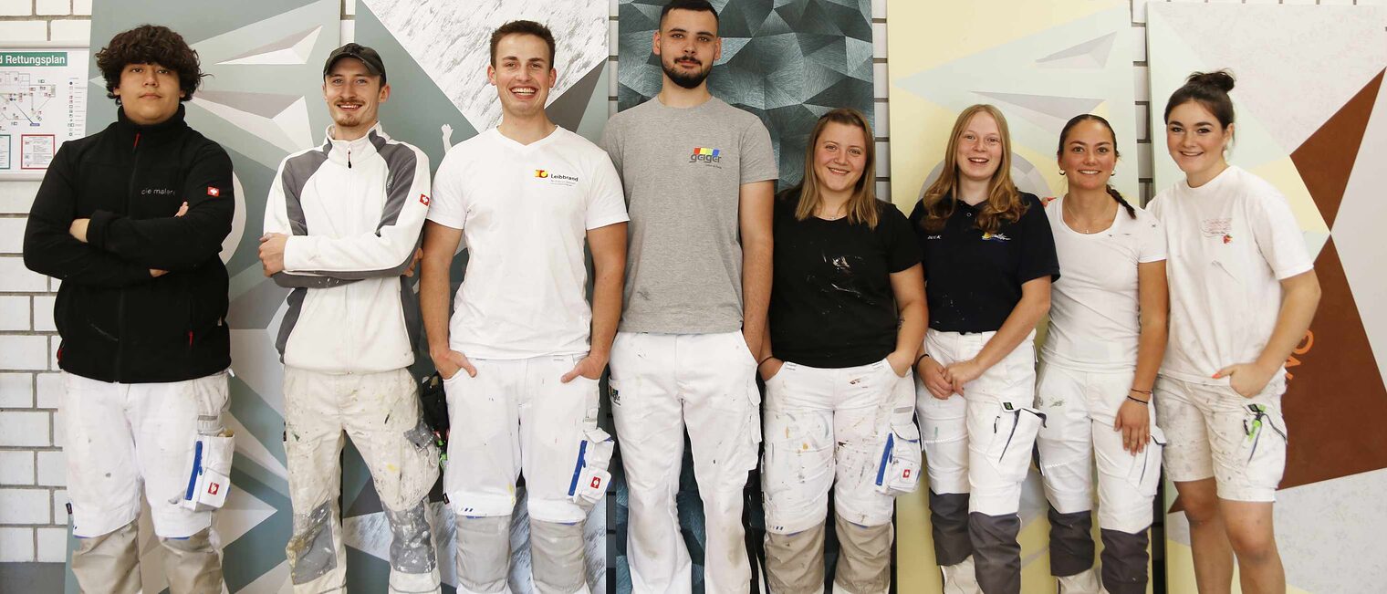8 junge Malerinnen und Maler, links die vier Jungs, rechts die vier Mädchen, alle in Maler-Kleidung vor bemalten Stellwänden, strahlen alle in die Kamera