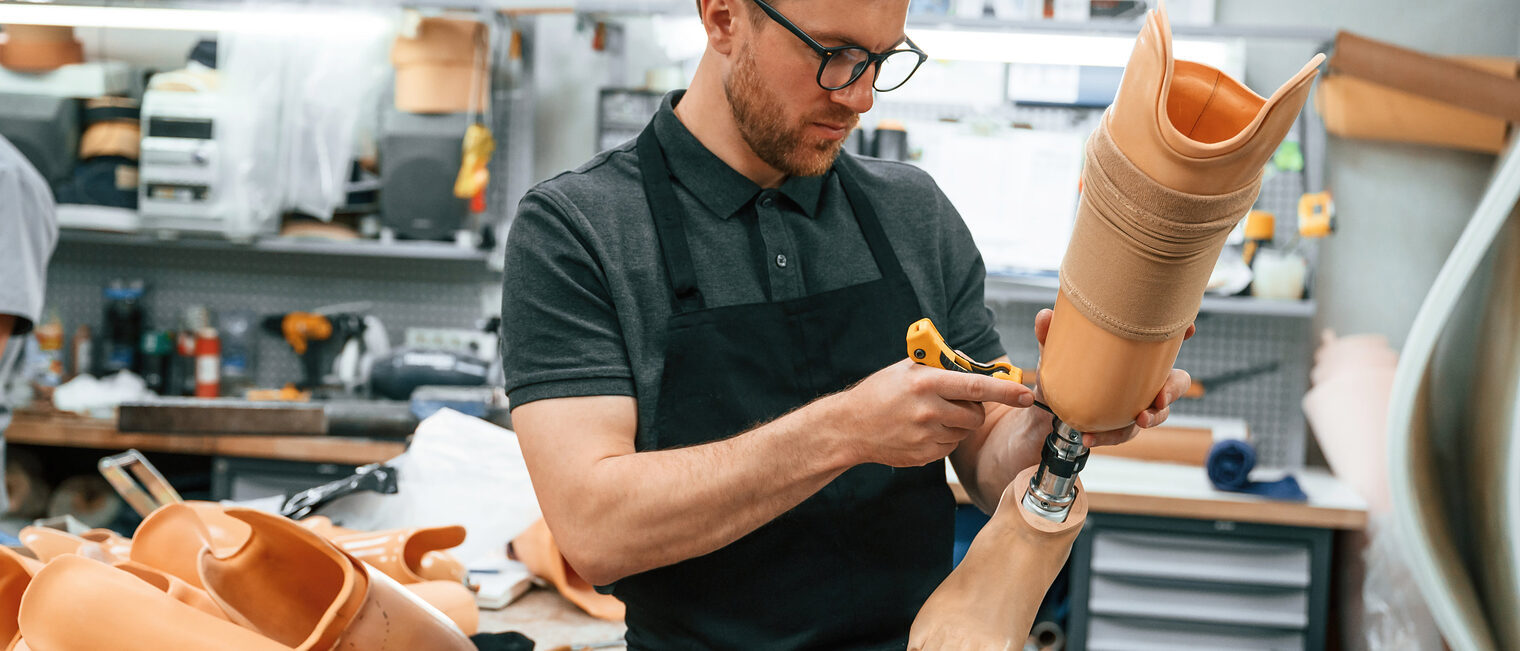 Orthopädietechnik-Mechaniker brauchen den elektronischen Berufsausweis eBA Mann mit kurzem Bart, Brille, dunklem Kragen-Shirt und schwarzer Schürze in einer Werkstatt für Orthopädietechnik, hält eine Beinprothese und schraubt an ihr