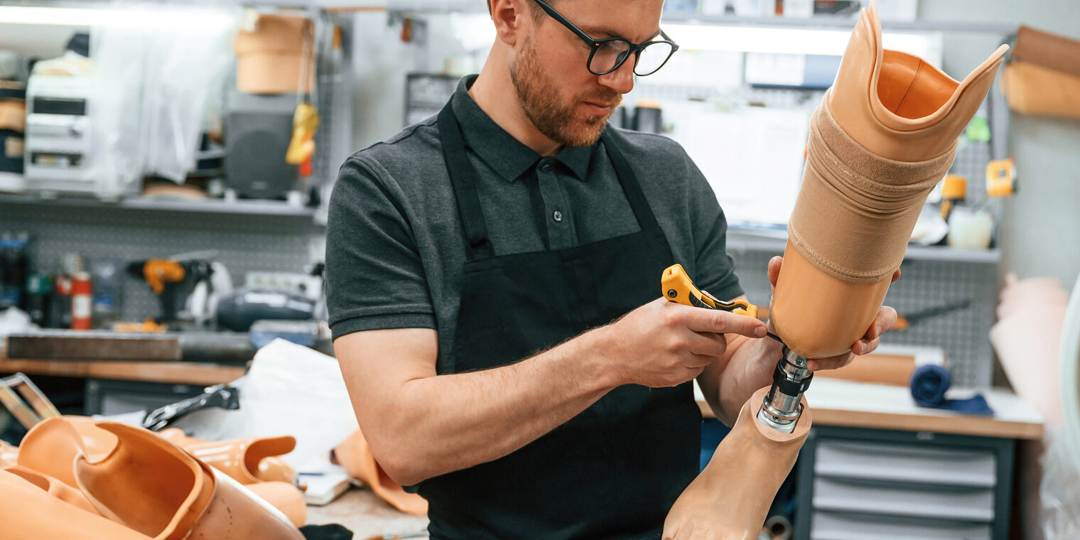 Orthopädietechnik-Mechaniker brauchen den elektronischen Berufsausweis eBA Mann mit kurzem Bart, Brille, dunklem Kragen-Shirt und schwarzer Schürze in einer Werkstatt für Orthopädietechnik, hält eine Beinprothese und schraubt an ihr