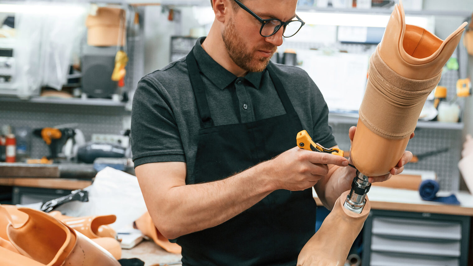 Mann mit kurzem Bart, Brille, dunklem Kragen-Shirt und schwarzer Schürze in einer Werkstatt für Orthopädietechnik, hält eine Beinprothese und schraubt an ihr