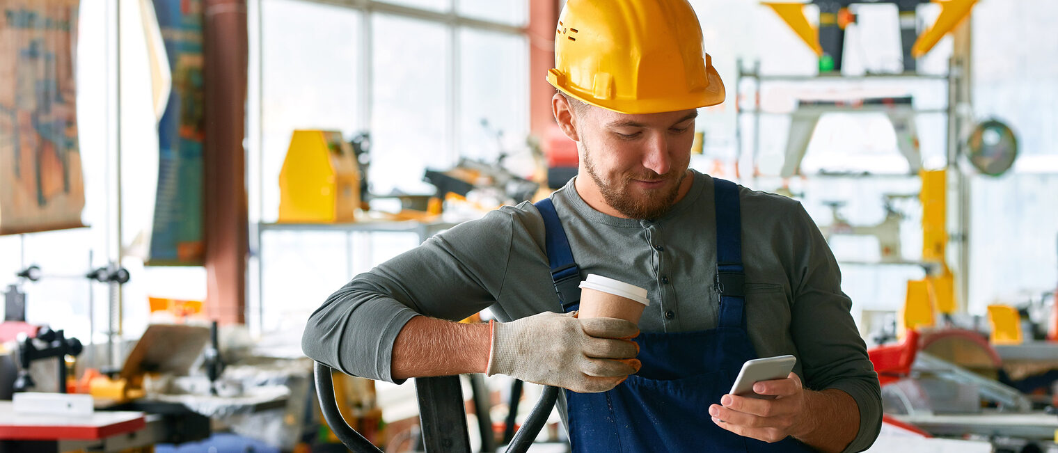 Handwerker mit blauer Latzhose und gelben Schutzhelm steht in seiner Werkstatt, in der rechten Hand ein Handy, auf das er schaut, die linke Hand hat einen Handschuh an und hält einen Papp-Kaffeebecher