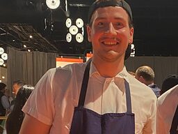 Ein Mann steht in Messehalle und bereite Backwaren zu, schaut in die Kamera. 