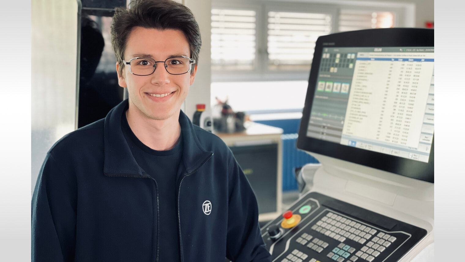 Junger Mann sitzt vor technischem Gerät und lächelt in die Kamera.