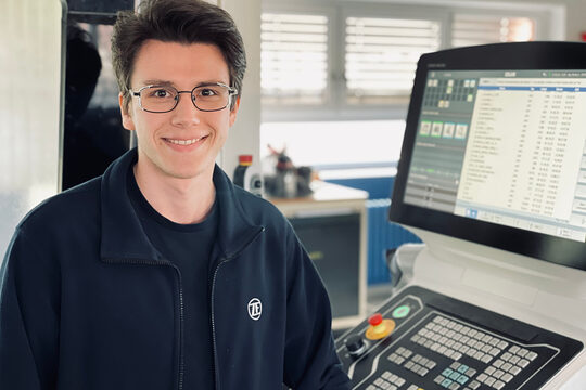 Meistergeschichte von Feinwerkmechaniker Floris Kalthoff Junger Mann sitzt vor technischem Gerät und lächelt in die Kamera.