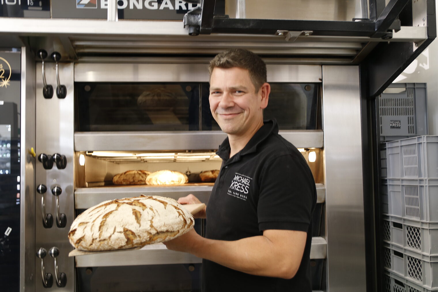 Mann steht vor Backofen und präsentiert ein gebackenes Brot, lächelt in die Kamera. 