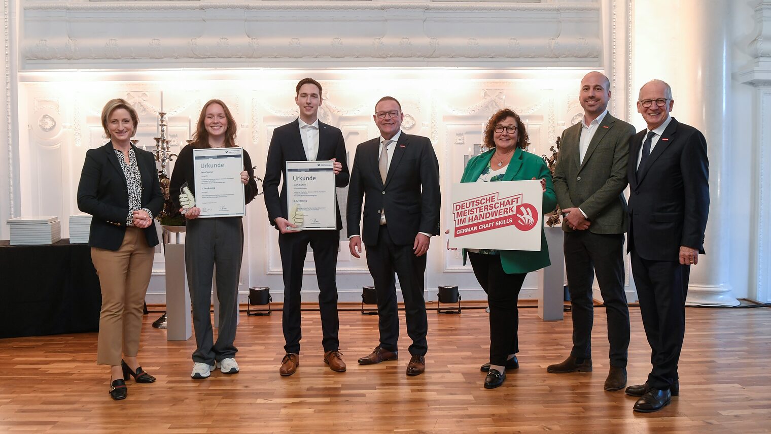 Sieben Personen in festlicher Kleidung stehen in einer Reihe und schauen in die Kamera. Zwei davon halten eine Urkunde. 