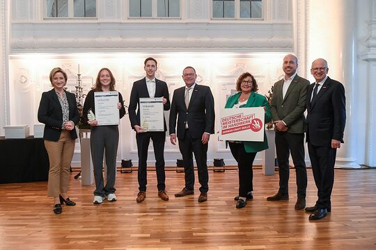 Sieben Personen in festlicher Kleidung stehen in einer Reihe und schauen in die Kamera. Zwei davon halten eine Urkunde. 