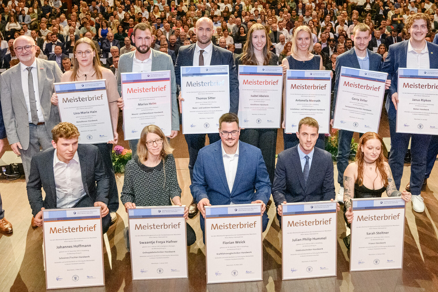 Gruppe von Menschen auf einer Bühne, 12 davon halten große Urkunden vor sich, im Hintergrund öffnet sich der Saal mit zahlreichen Besuchern