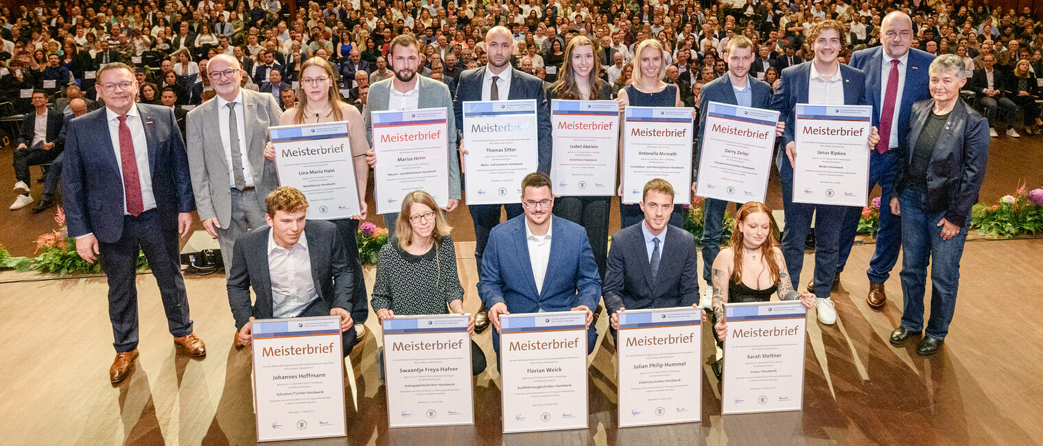 Gruppe von Menschen auf einer Bühne, 12 davon halten große Urkunden vor sich, im Hintergrund öffnet sich der Saal mit zahlreichen Besuchern