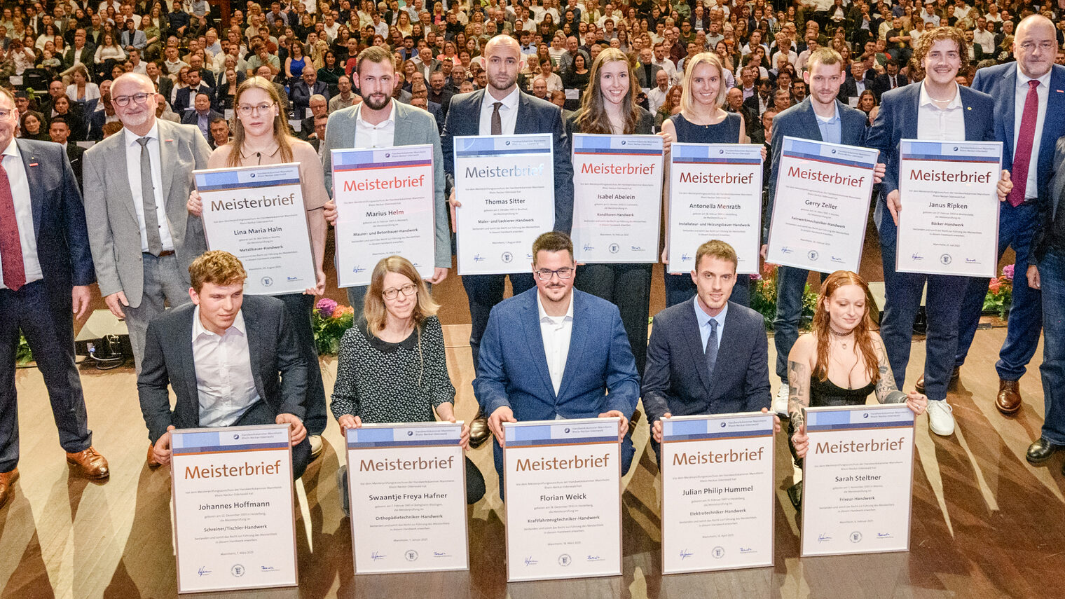 Gruppe von Menschen auf einer Bühne, 12 davon halten große Urkunden vor sich, im Hintergrund öffnet sich der Saal mit zahlreichen Besuchern