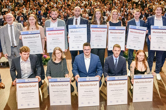 Gruppe von Menschen auf einer Bühne, 12 davon halten große Urkunden vor sich, im Hintergrund öffnet sich der Saal mit zahlreichen Besuchern