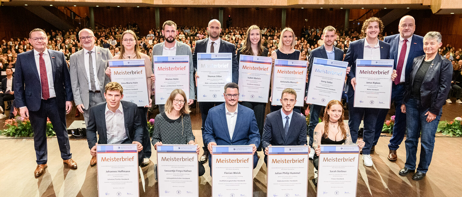 Gruppe von Menschen auf einer Bühne, 12 davon halten große Urkunden vor sich, im Hintergrund öffnet sich der Saal mit zahlreichen Besuchern