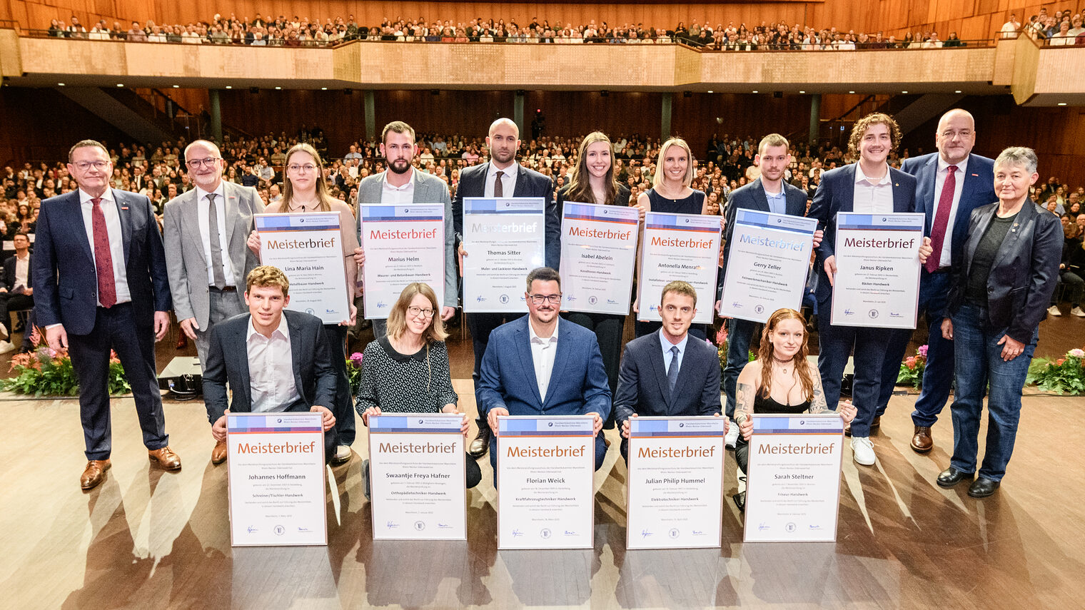 Gruppe von Menschen auf einer Bühne, 12 davon halten große Urkunden vor sich, im Hintergrund öffnet sich der Saal mit zahlreichen Besuchern