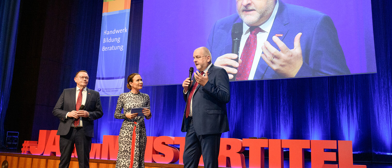 Drei Personen stehen auf der Bühne und ein Mann spricht in ein Mikrofon