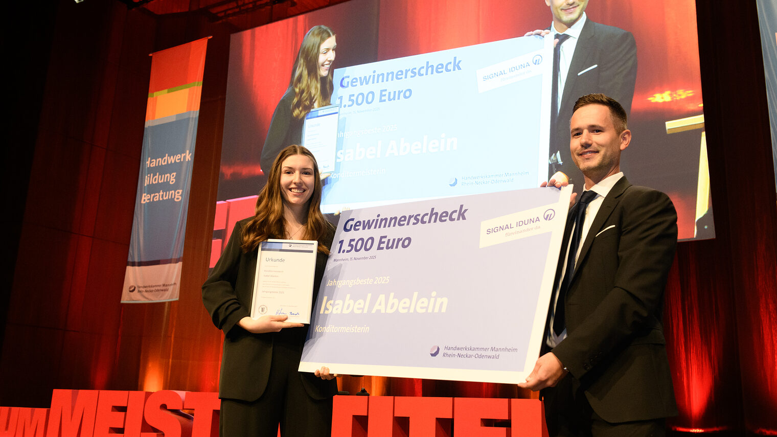 Eine Frau und ein Mann stehen auf einer Bühne und halten einen großen Gewinnercheck in der Mitte, lächeln in die Kamera.