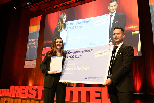 Eine Frau und ein Mann stehen auf einer Bühne und halten einen großen Gewinnercheck in der Mitte, lächeln in die Kamera.