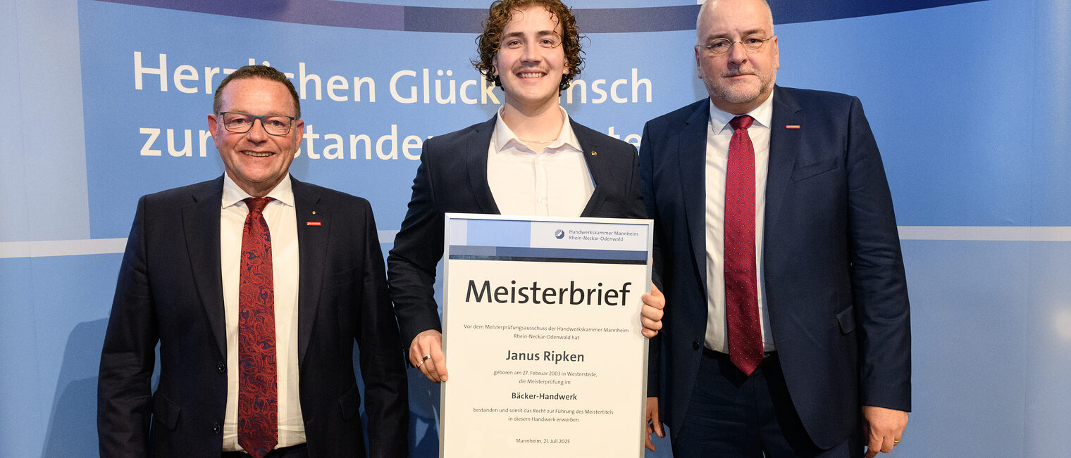 Drei Personen stehen vor einer Wand, einer hält seinen gerahmten Meisterbrief