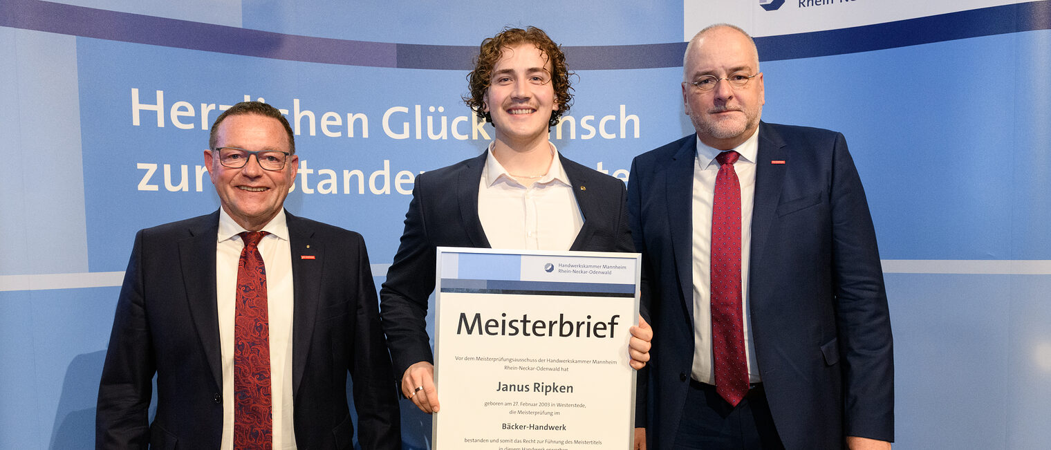 Drei Personen stehen vor einer Wand, einer hält seinen gerahmten Meisterbrief