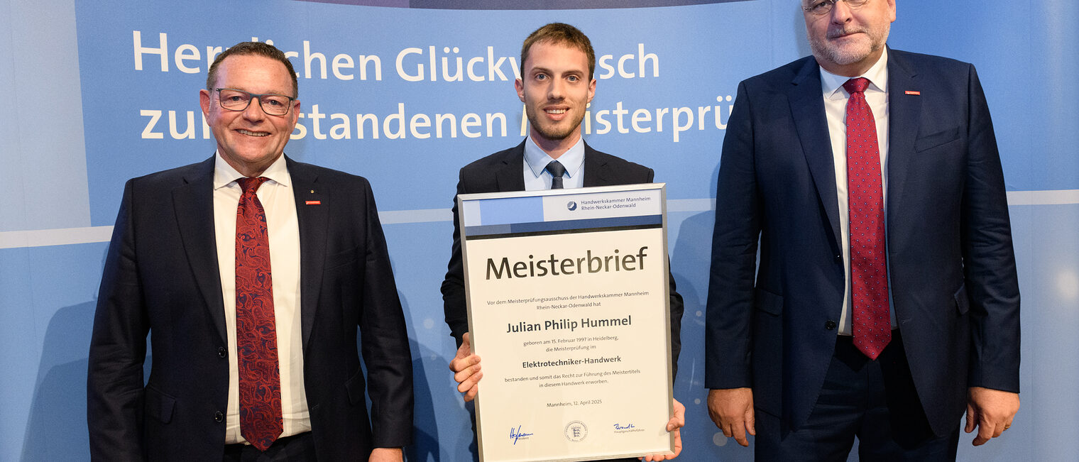 Drei Personen stehen vor einer Wand, einer hält seinen gerahmten Meisterbrief