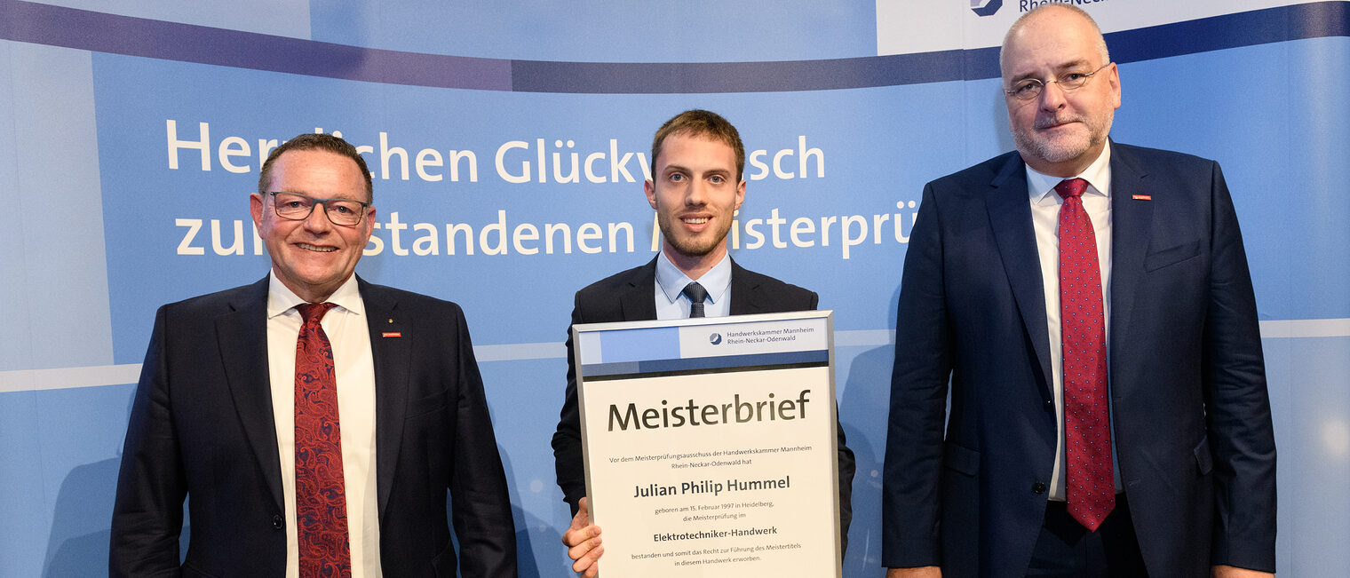 Drei Personen stehen vor einer Wand, einer hält seinen gerahmten Meisterbrief