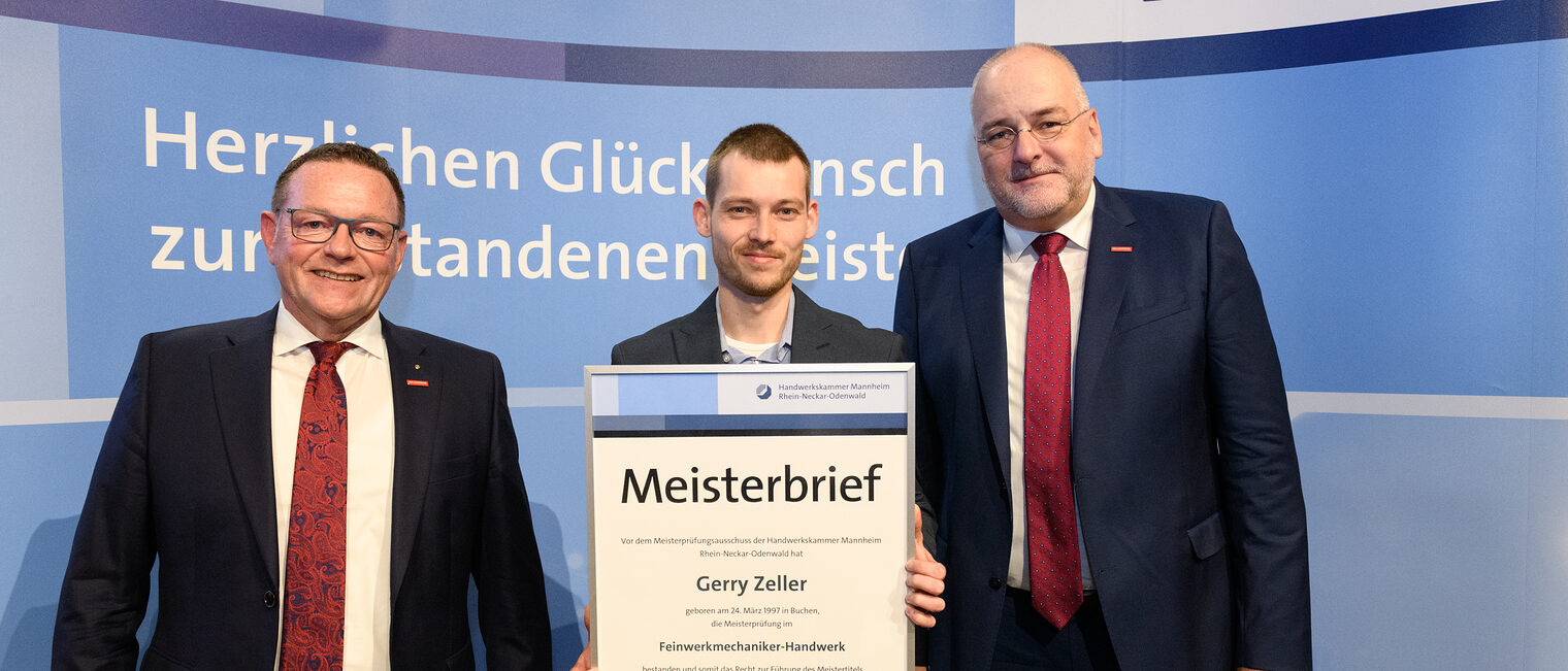 Drei Personen stehen vor einer Wand, einer hält seinen gerahmten Meisterbrief