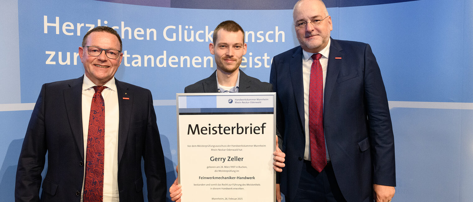 Drei Personen stehen vor einer Wand, einer hält seinen gerahmten Meisterbrief