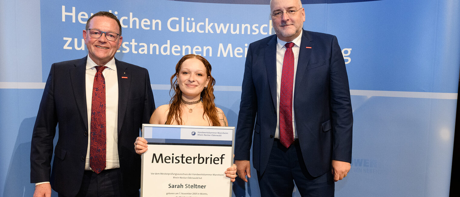 Drei Personen stehen vor einer Wand und eine hält einen gerahmten Meisterbrief in der Hand