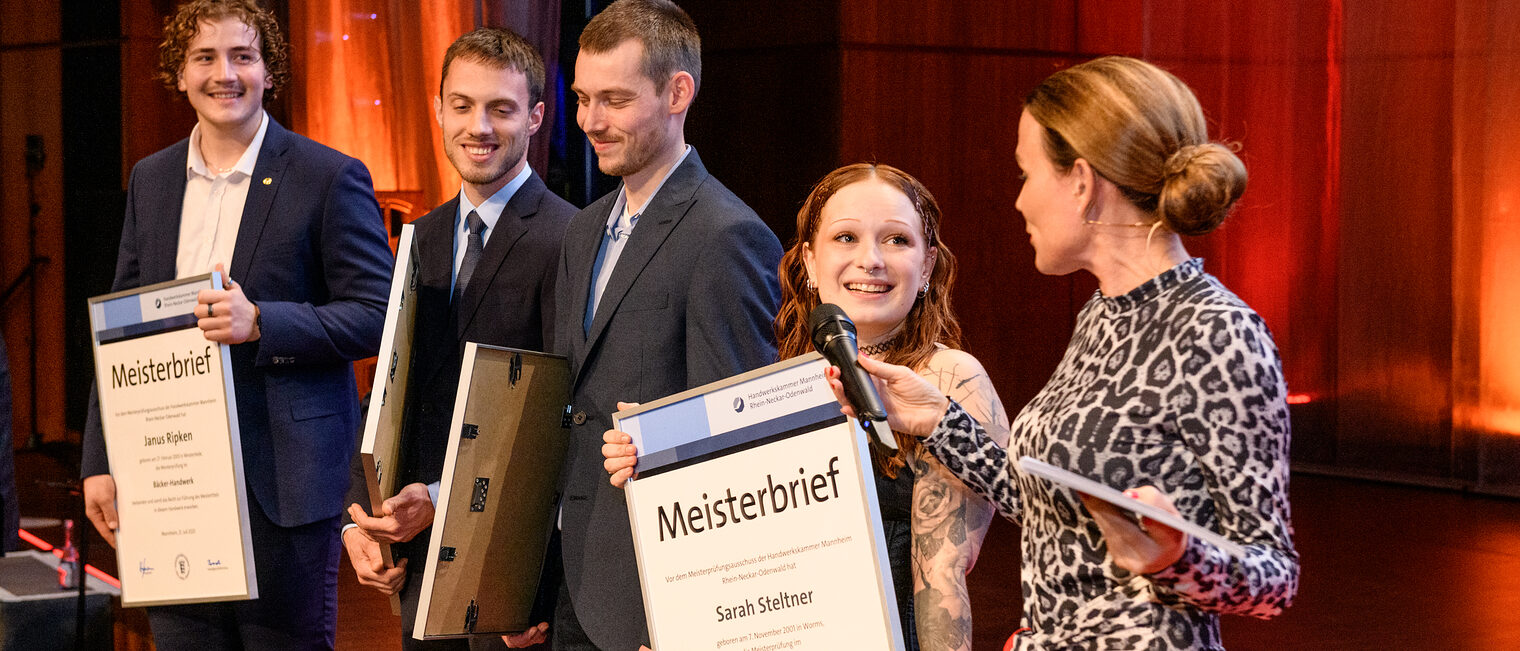 Auf der Bühne stehen Personen, und die Moderatorin stellt Fragen mit einem Handmikrofon