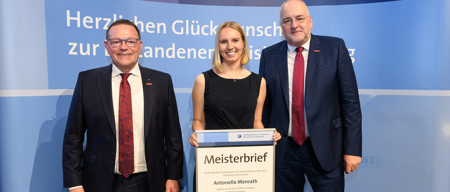 Drei Personen stehen vor einer Wand und eine hält einen gerahmten Meisterbrief in den Händen