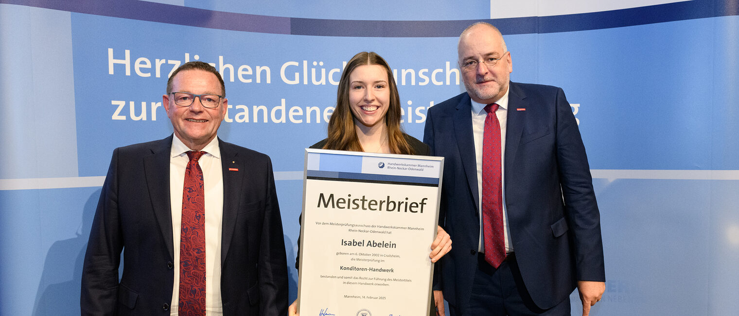 Drei Personen stehen vor einer Wand und eine hält einen gerahmten Meisterbrief in den Händen