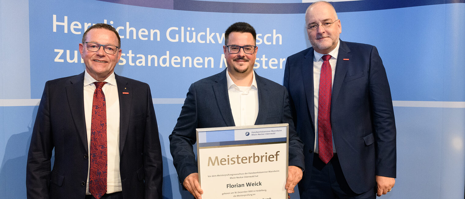 Drei Personen stehen vor einer Wand und eine hält einen gerahmten Meisterbrief in den Händen