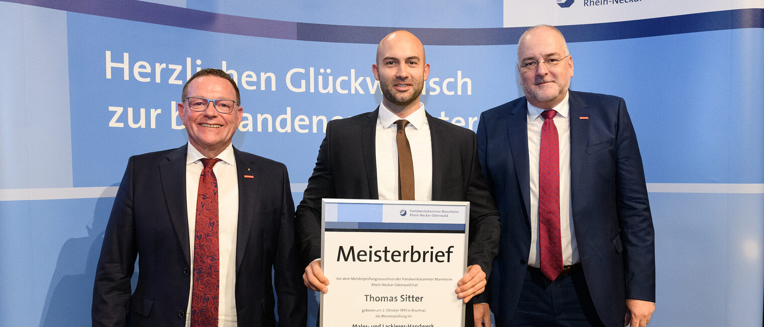 Drei Personen stehen wir einer Wand, einer hält einen gerahmten Meisterbrief in den Händen