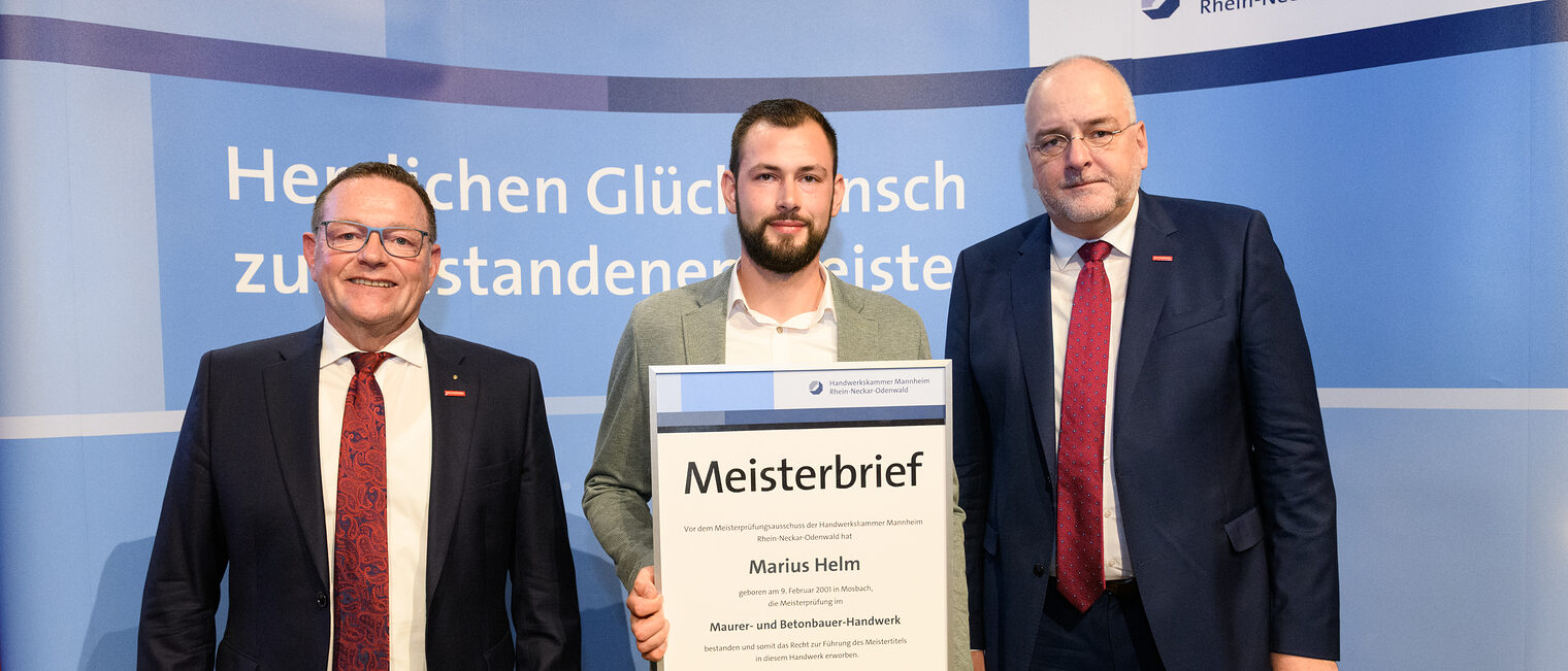 Drei Personen stehen wir einer Wand, einer hält einen gerahmten Meisterbrief in den Händen