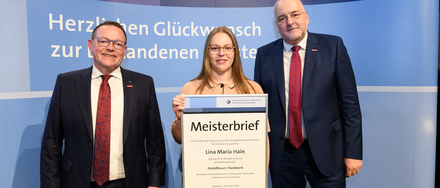 Drei Personen stehen wir einer Wand, einer hält einen gerahmten Meisterbrief in den Händen