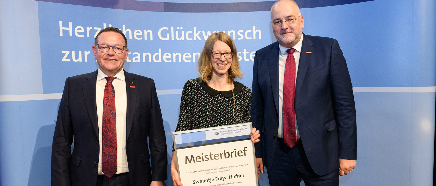 Drei Personen stehen wir einer Wand, einer hält einen gerahmten Meisterbrief in den Händen