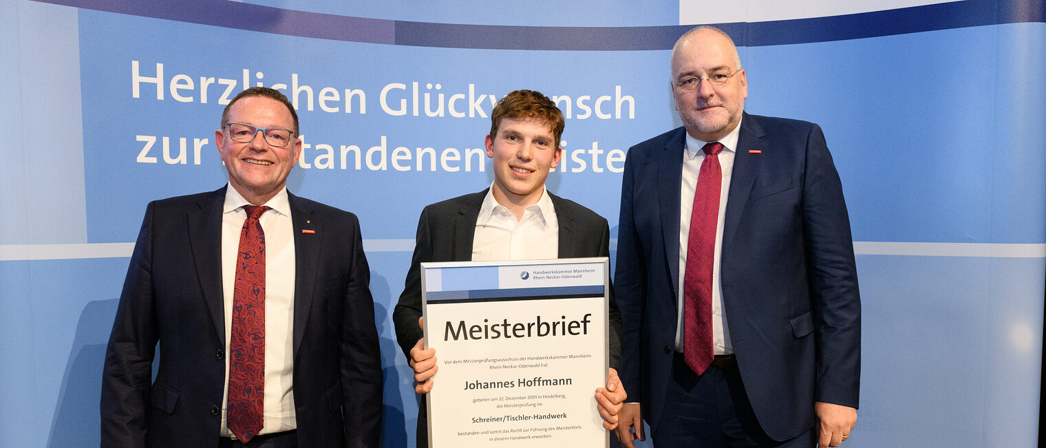 Drei Personen stehen wir einer Wand, einer hält einen gerahmten Meisterbrief in den Händen