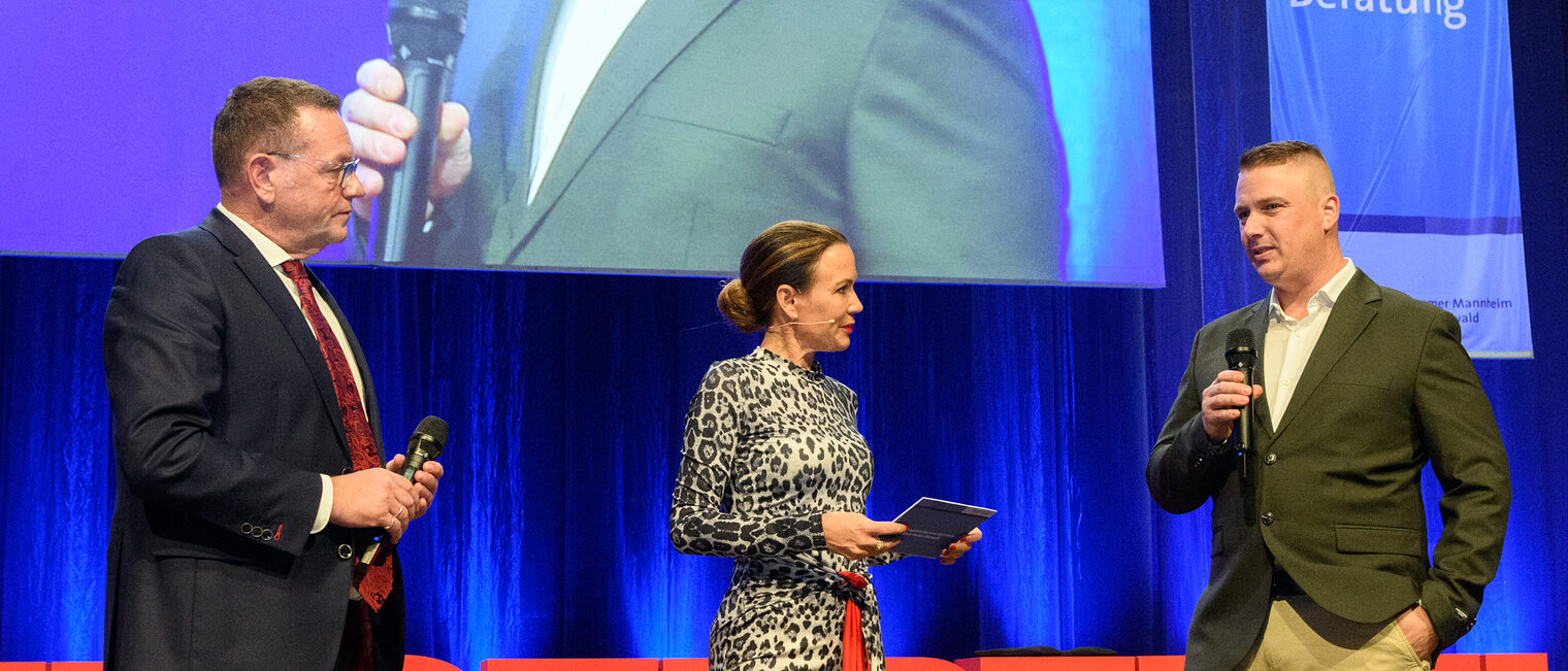 Drei Personen stehen auf der Bühne, die zwei Männer halten ein Handmikrofon