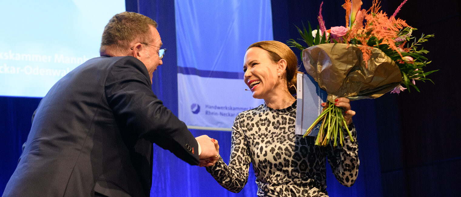 Zwei Personen stehen auf einer Bühne, der Mann schüttelt der Frau die Hand und überreicht ihr einen Strauß Blumen